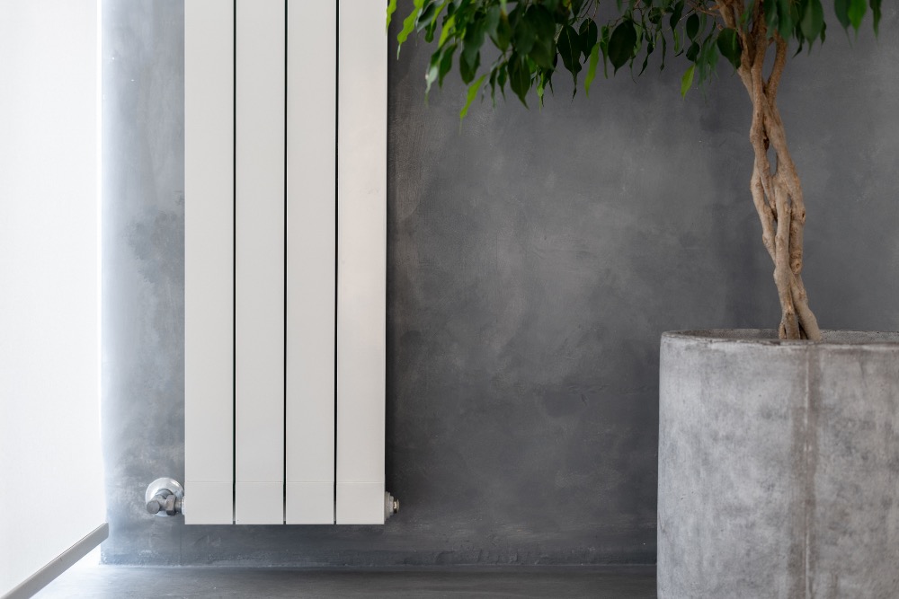 Close up of white vertical radiator on a grey concrete wall next to small tree plant