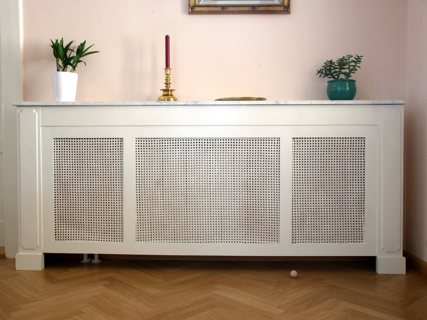 White radiator cover with perforated panels, topped with two potted plants and a brass candlestick.