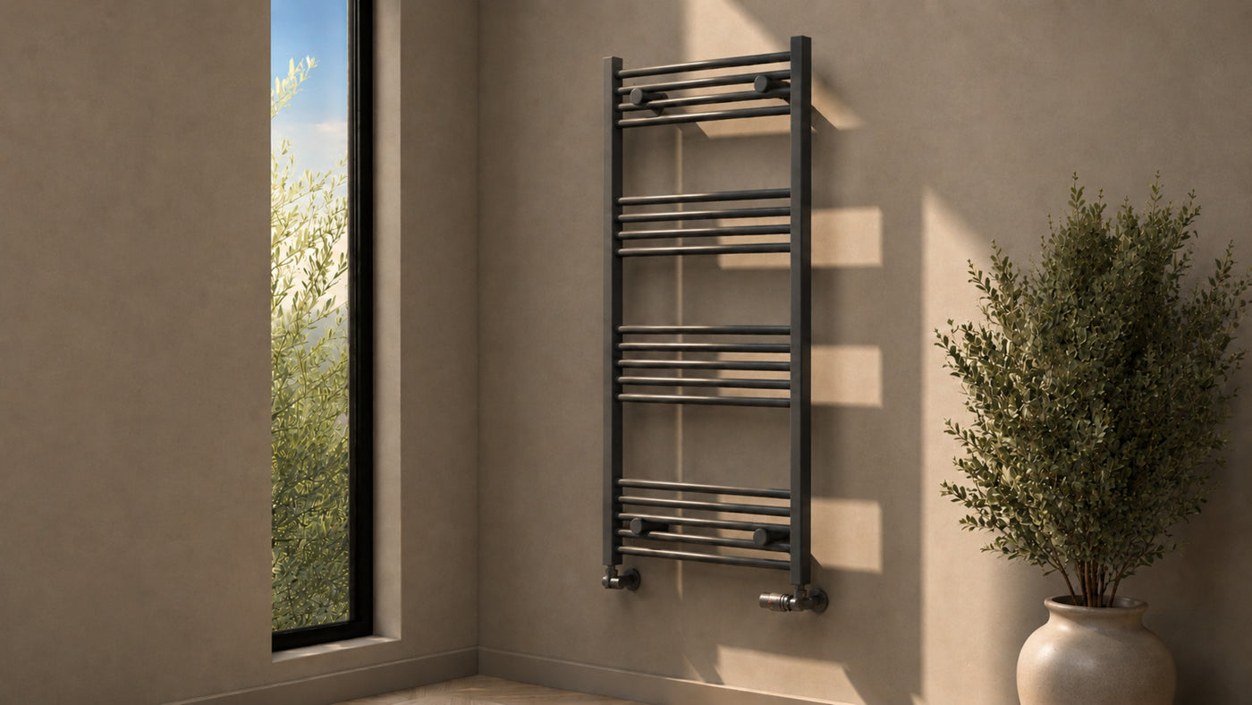 Wide view of an anthracite heated towel rail with matching corner valves in a sunlit bathroom beside a tall window and potted plant.