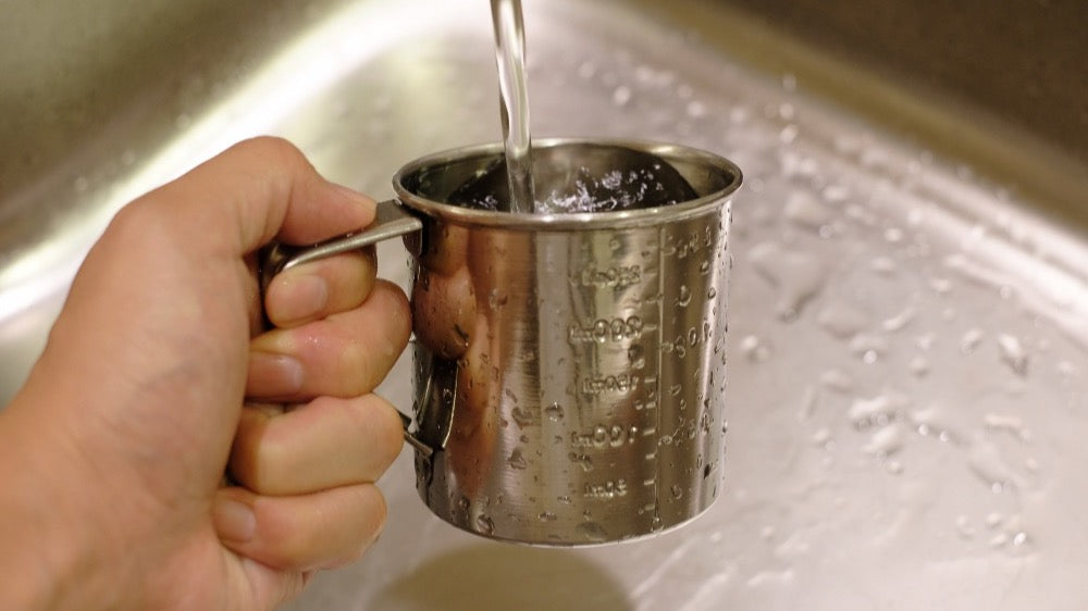 Hand holding a stainless steel measuring jug under a running kitchen tap to measure water flow rate in litres per minute.
