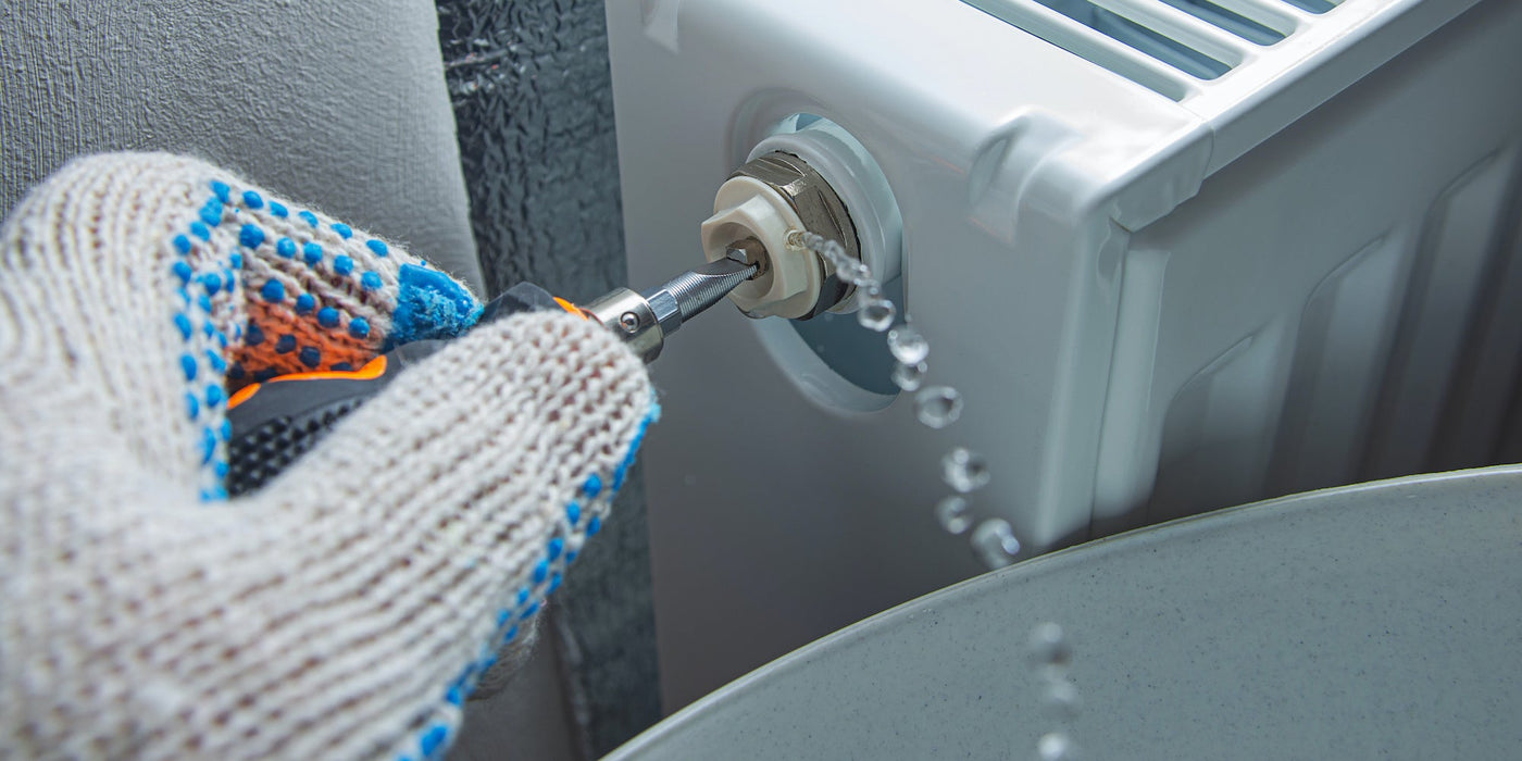 Gloved hand using a radiator bleed key to bleed a panel radiator as water drains into a bucket.