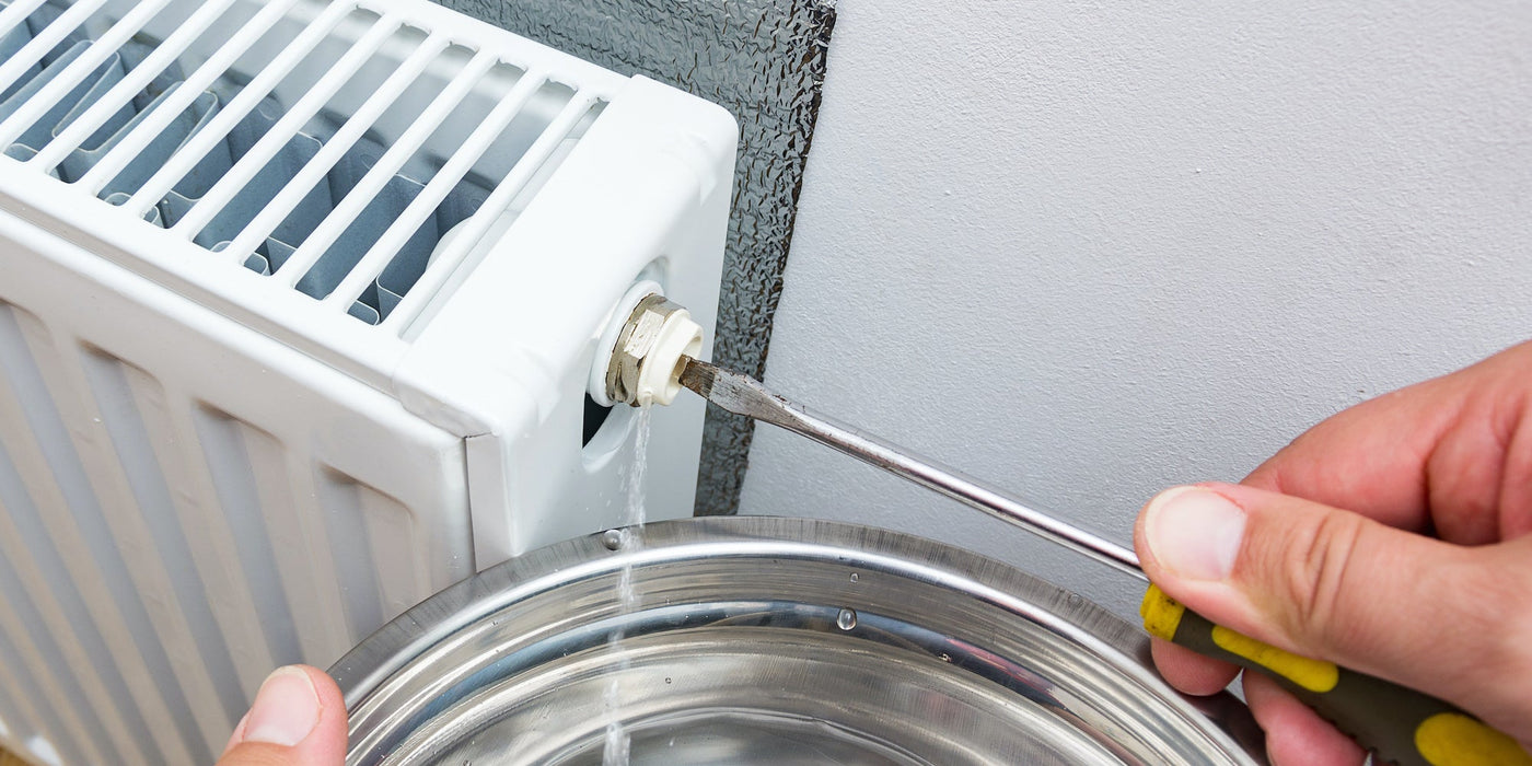 Hand using a flathead screwdriver to open a radiator bleed valve while water drains into a metal bowl.