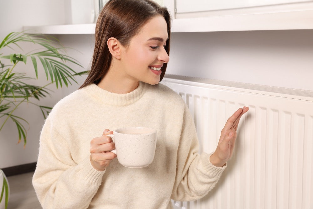 Woman holding a mug while checking the temperature of a panel radiator by placing her hand against the front, indicating a warm and functioning central heating system.